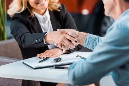 A professional woman and a man shaking hands across a desk with documents and car keys, symbolizing an agreement or legal consultation