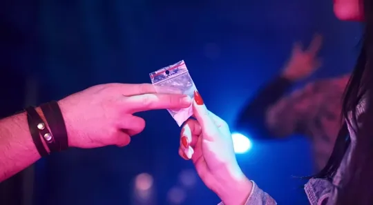 An image of two people exchanging a small plastic bag, with flashing police lights in a dark, blurred background.
