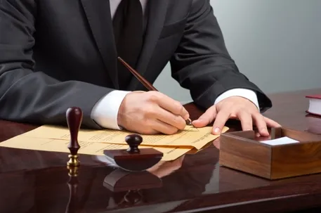 A man wearing a formal suit, writing at a table, with a stamp beside him.