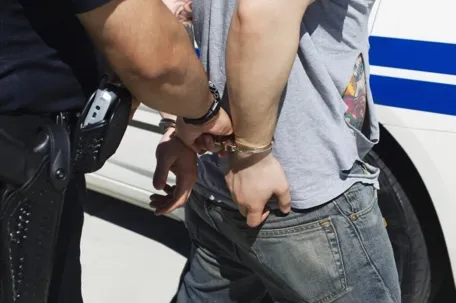 An image of a man being arrested by the police, with his hands handcuffed, being escorted to a patrol car.